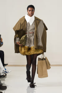 Model walking a runway in a layered, brown ensemble with a oversized white collar and fur-trimmed hem, carrying shopping bags.