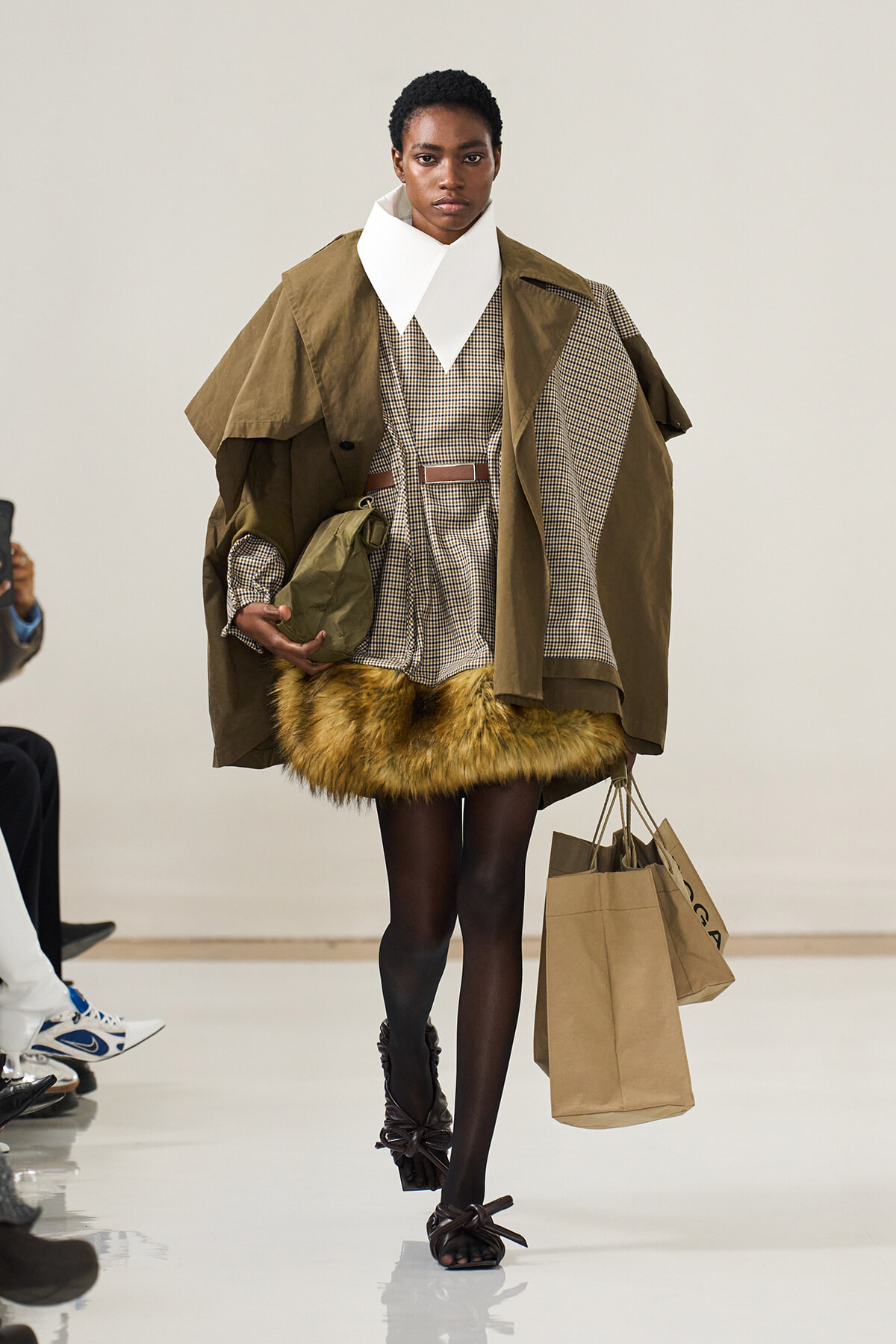 Model walking a runway in a layered, brown ensemble with a oversized white collar and fur-trimmed hem, carrying shopping bags.