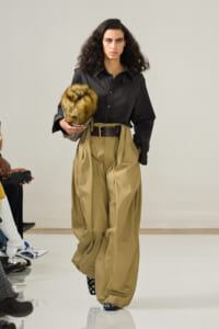 Model in a black blouse and oversized tan pants walks a fashion runway, holding a fur hat.