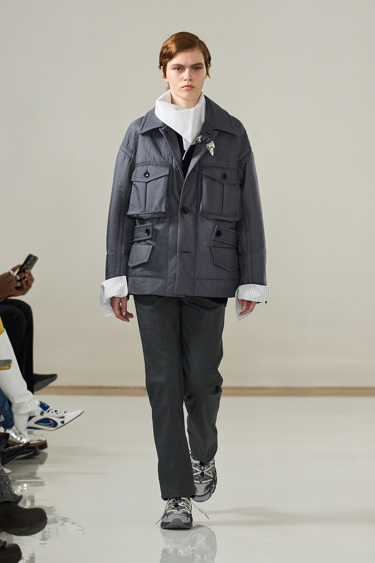 Model on runway wearing a oversized gray utility jacket with multiple pockets, dark gray trousers, and patterned sneakers; white check shirt cuffs peeking out.
