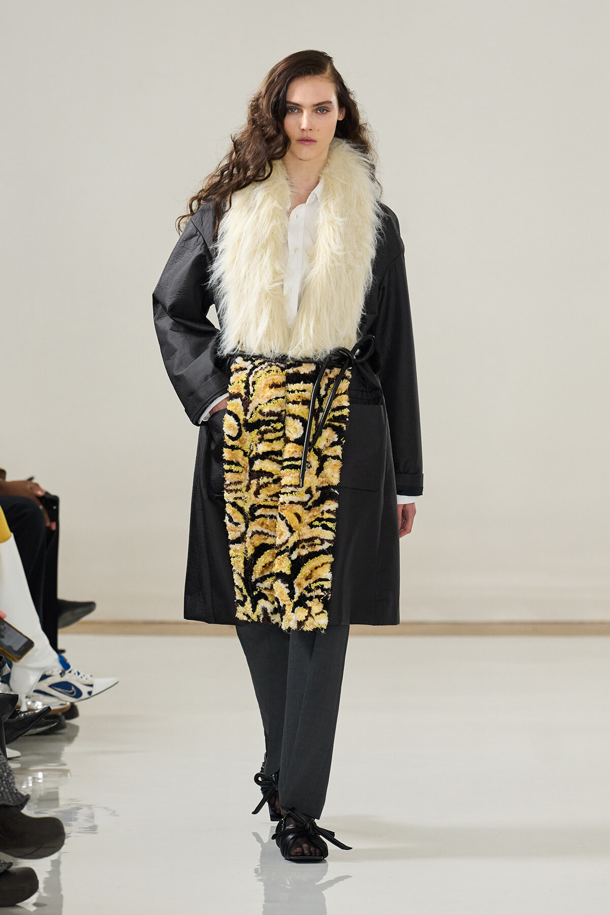 Model on a runway wearing an oversized black coat with a cream faux-fur scarf and a tiger-print belt over grey trousers.