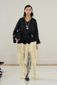 Model on a runway wearing a black silky blouse and a cream fringe skirt with black tights and heels, accessorized with a chunky silver necklace and clutch.