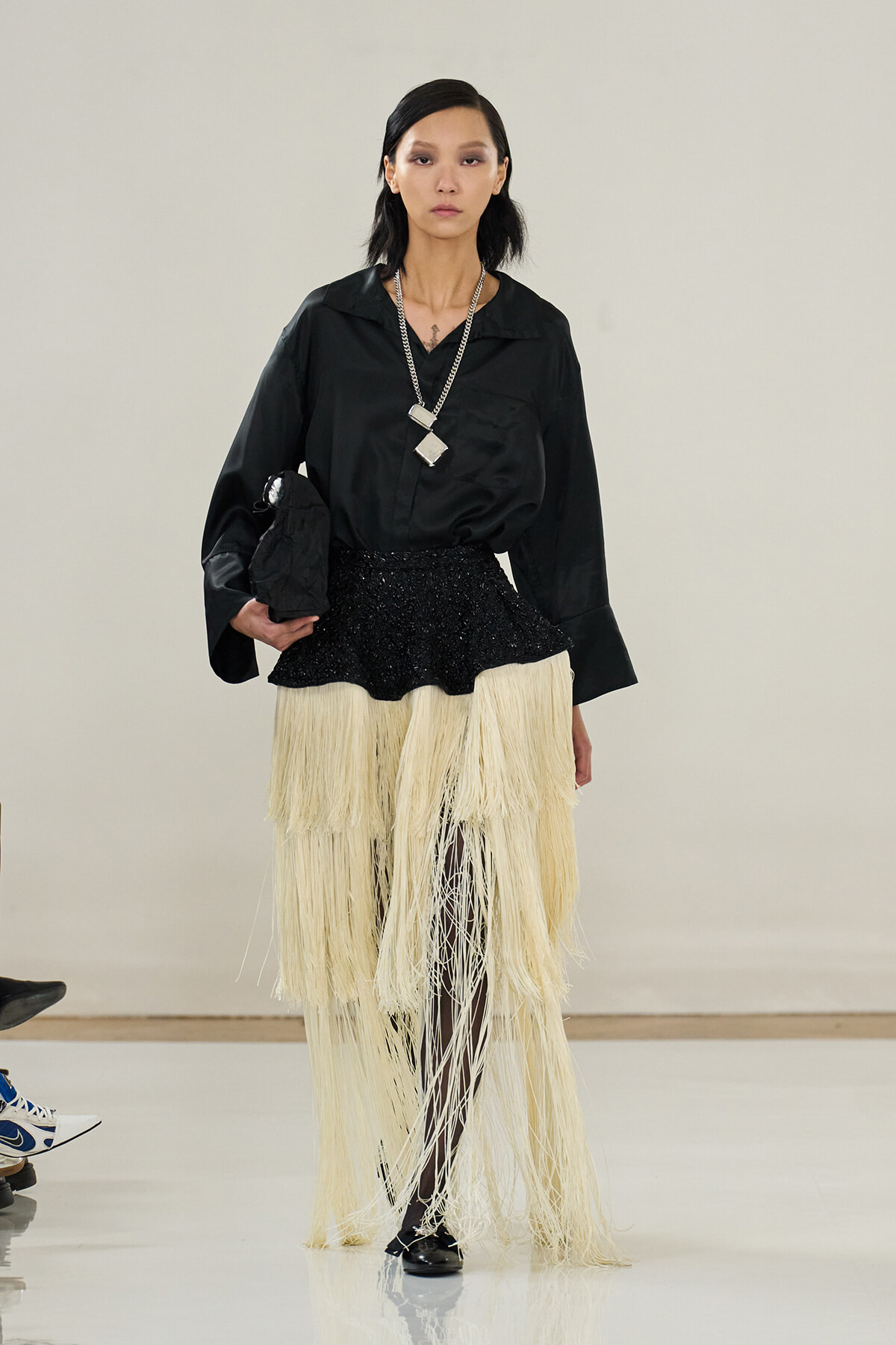 Model on a runway wearing a black silky blouse and a cream fringe skirt with black tights and heels, accessorized with a chunky silver necklace and clutch.