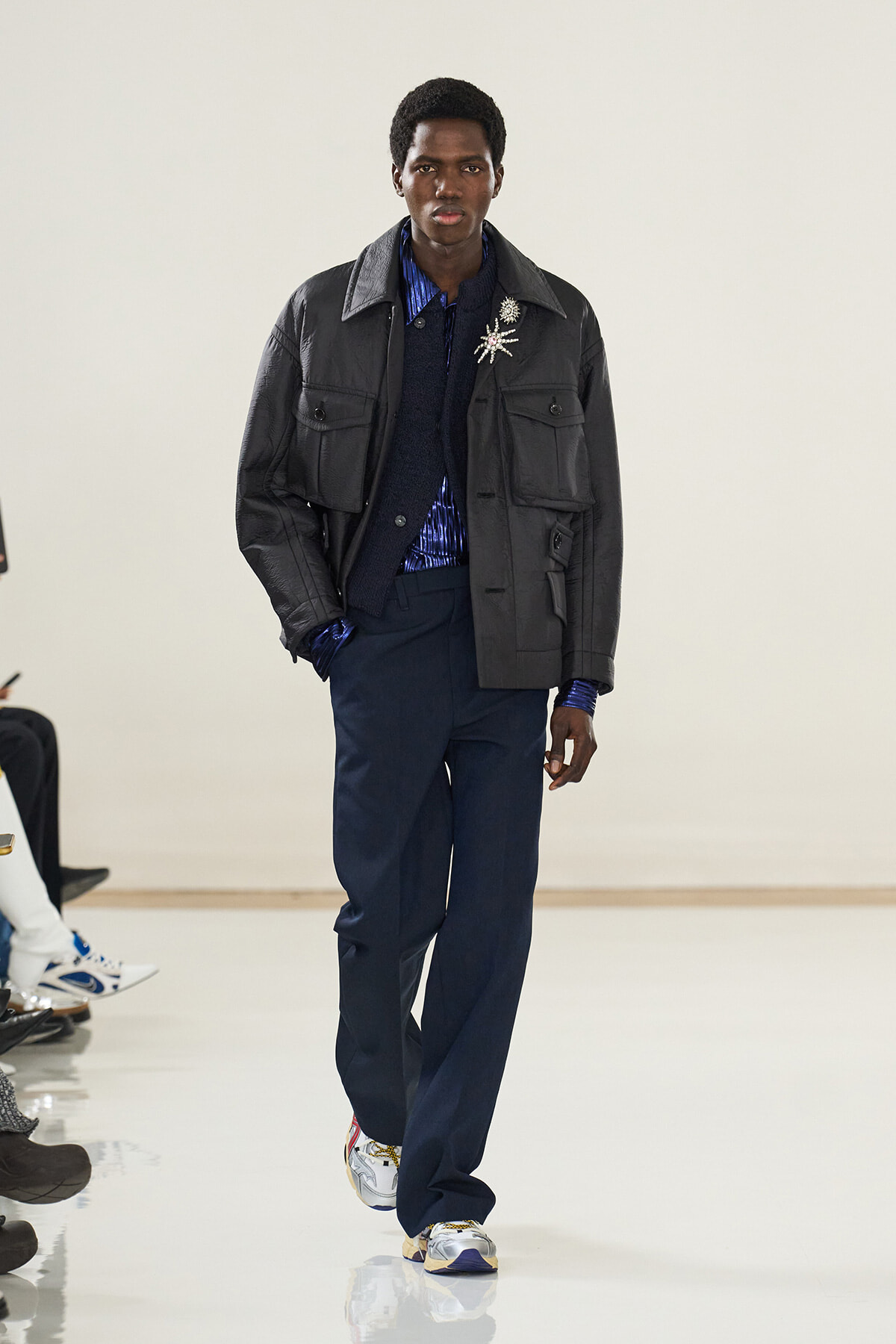 Male model walking the runway in a black quilted jacket over a blue striped shirt and navy trousers, white sneakers visible at the feet, neutral backdrop.