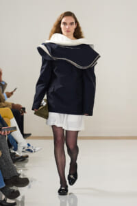 Runway model wearing an oversized navy coat with a large white scarf collar, white skirt, black tights, and black loafers, holding a small olive bag.