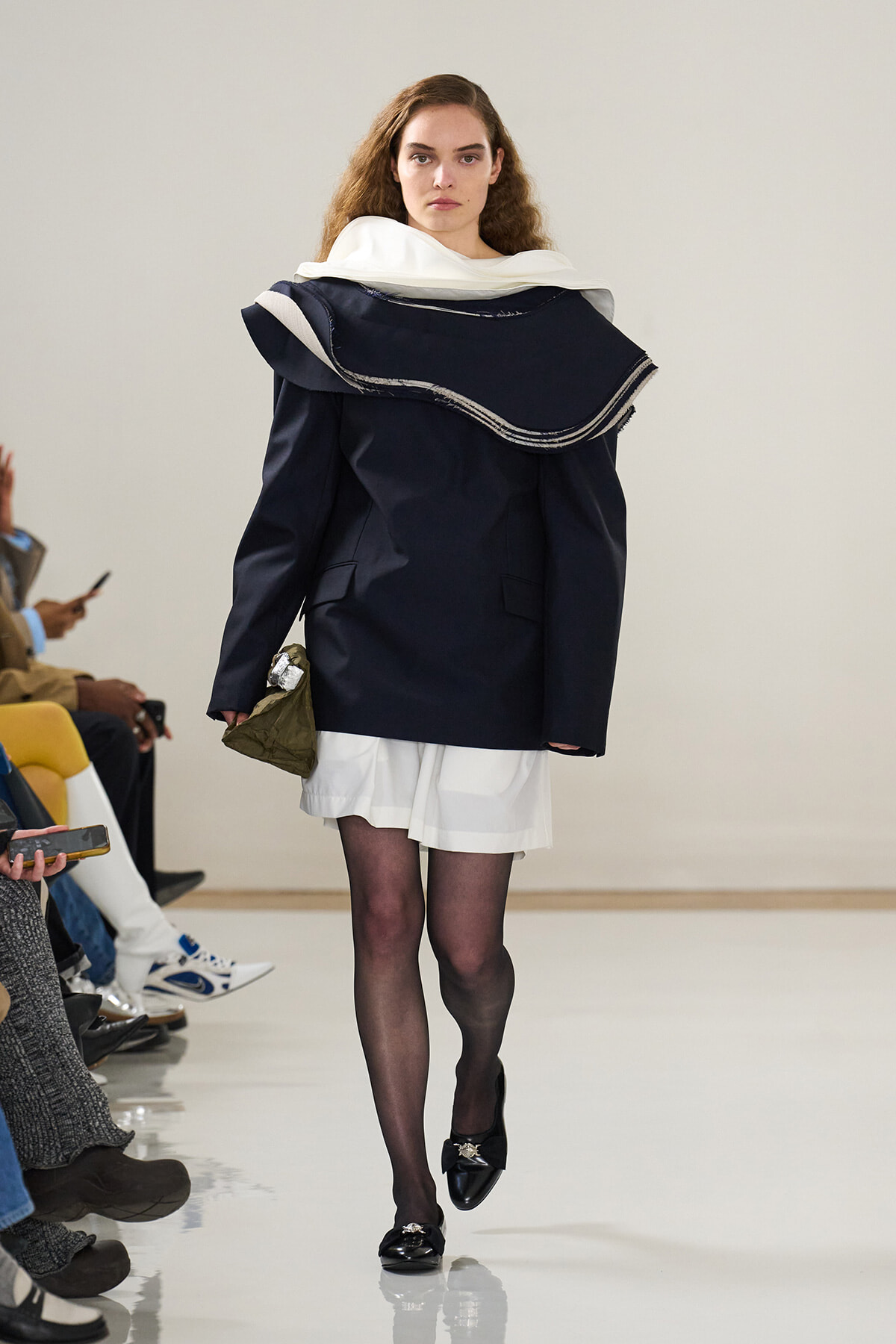 Runway model wearing an oversized navy coat with a large white scarf collar, white skirt, black tights, and black loafers, holding a small olive bag.