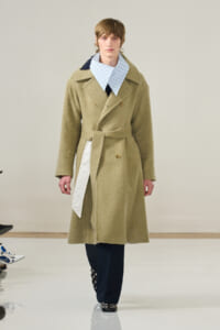 Male model on a runway wearing a beige belted wool coat with wide lapels, paired with a light blue checkered shirt and dark trousers with studs at the hem.