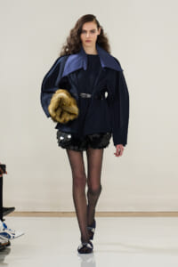 Model on a runway wearing a dark navy oversized coat with a large blue collar, cinched at the waist, paired with black sequined shorts and sheer tights, holding a fur stole.
