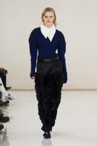 Female model walks the runway in a navy ribbed sweater with a large white collar, black belt, and voluminous ruffled black pants, audience visible to the left.