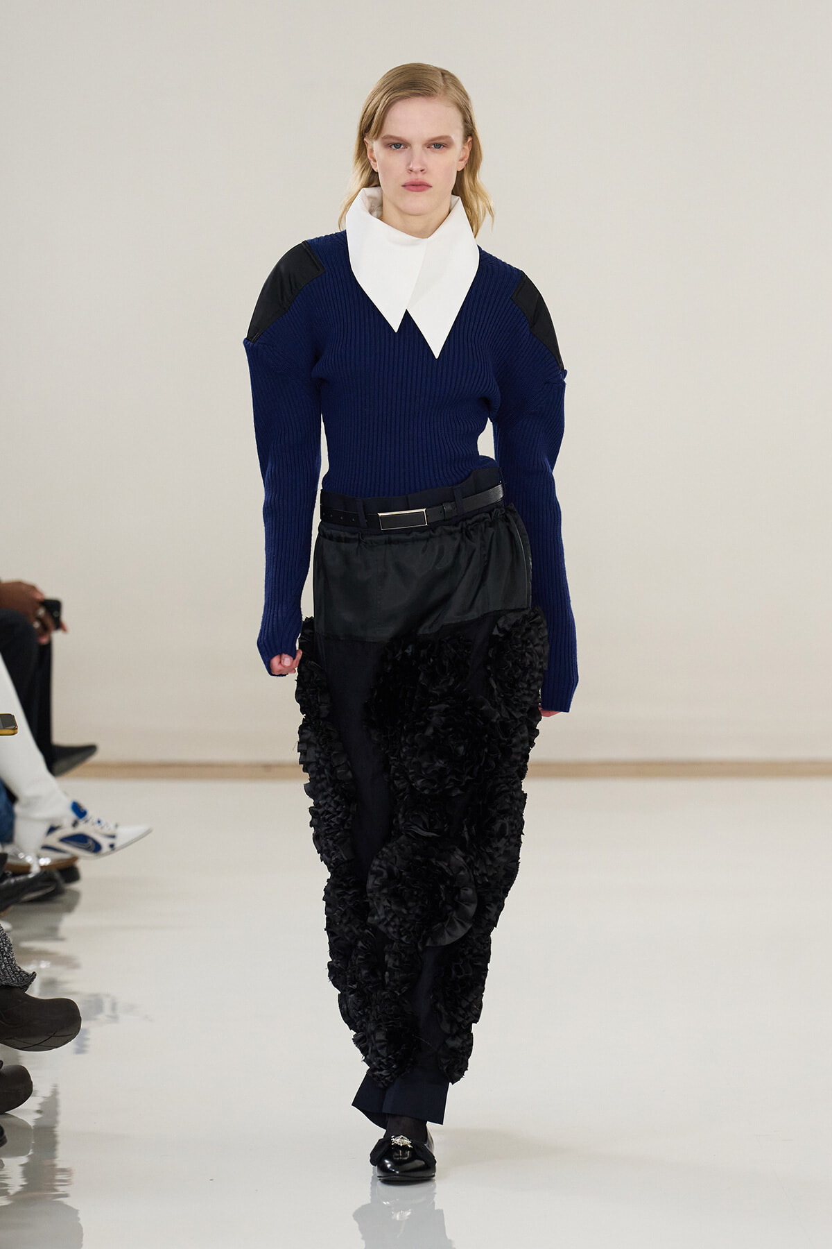 Female model walks the runway in a navy ribbed sweater with a large white collar, black belt, and voluminous ruffled black pants, audience visible to the left.