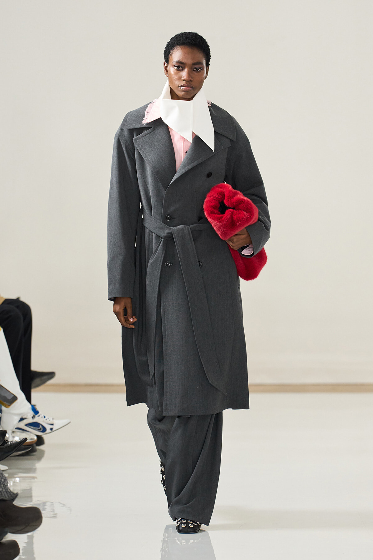 Model walks a runway in a long gray trench coat, with a white oversized collar and pink blouse, holding a red fur heart clutch.
