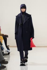 Model on a runway wearing a navy oversized coat with a hooded top, black gloves, and a bright red bag; distinctive fur-trimmed bow shoes are visible at the feet.