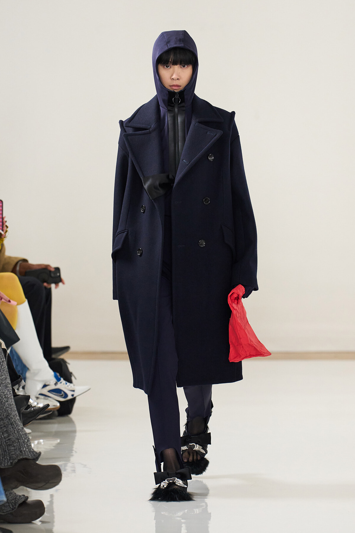 Model on a runway wearing a navy oversized coat with a hooded top, black gloves, and a bright red bag; distinctive fur-trimmed bow shoes are visible at the feet.