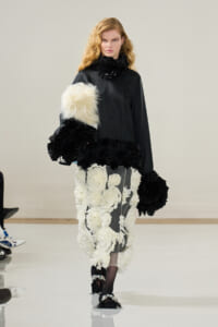 Model walking a runway in a black top with a voluminous white floral-appliqué skirt and large black fur accents on sleeves and hem.