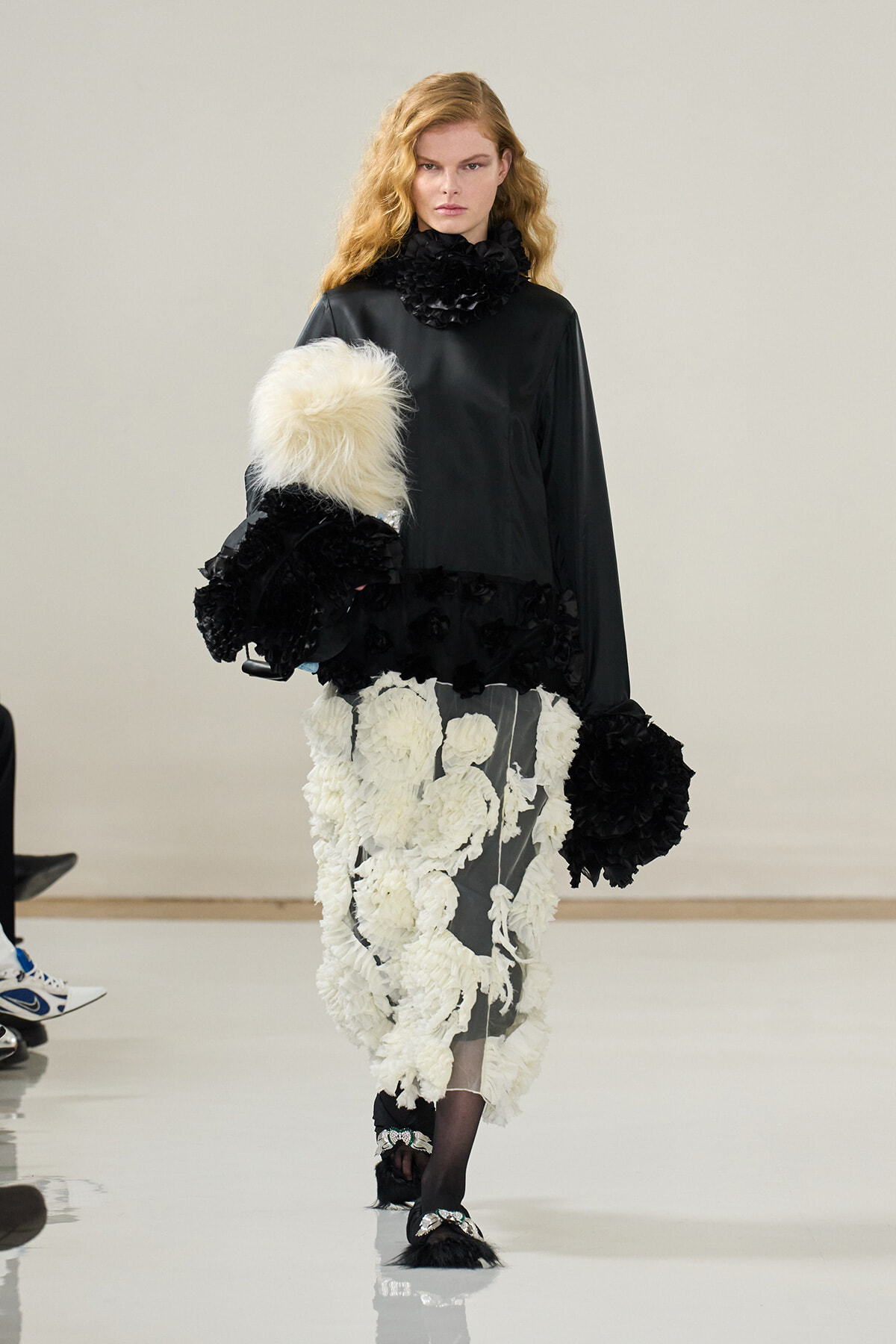 Model walking a runway in a black top with a voluminous white floral-appliqué skirt and large black fur accents on sleeves and hem.