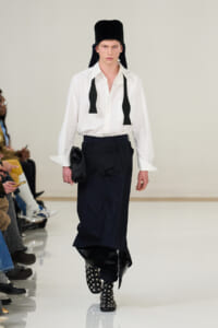 Male model walks the runway in a white button-down shirt, a dark maxi skirt, and black studded boots, with a tall black headpiece and hanging ties.