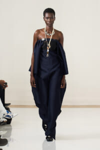 Model on a runway wearing a navy off-shoulder cape jumpsuit with layered pearl necklaces and black shoes, pose-oriented.