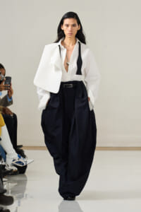 Model walking a runway in a white oversized blouse and voluminous black pants, accessorized with a silver chain necklace and belt.