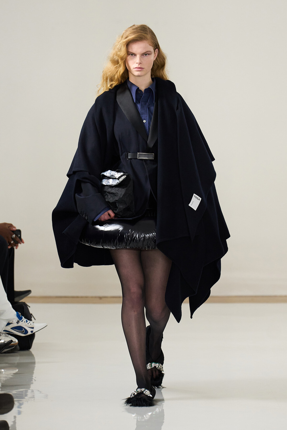 Model walks the runway in a navy cape over a blue shirt, belted at the waist, with a shiny black mini skirt and embellished feathered sandals.