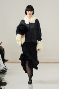 Runway model in a black satin outfit with white ruffled collar and cuffs, carrying a black fur accessory and wearing black heels.