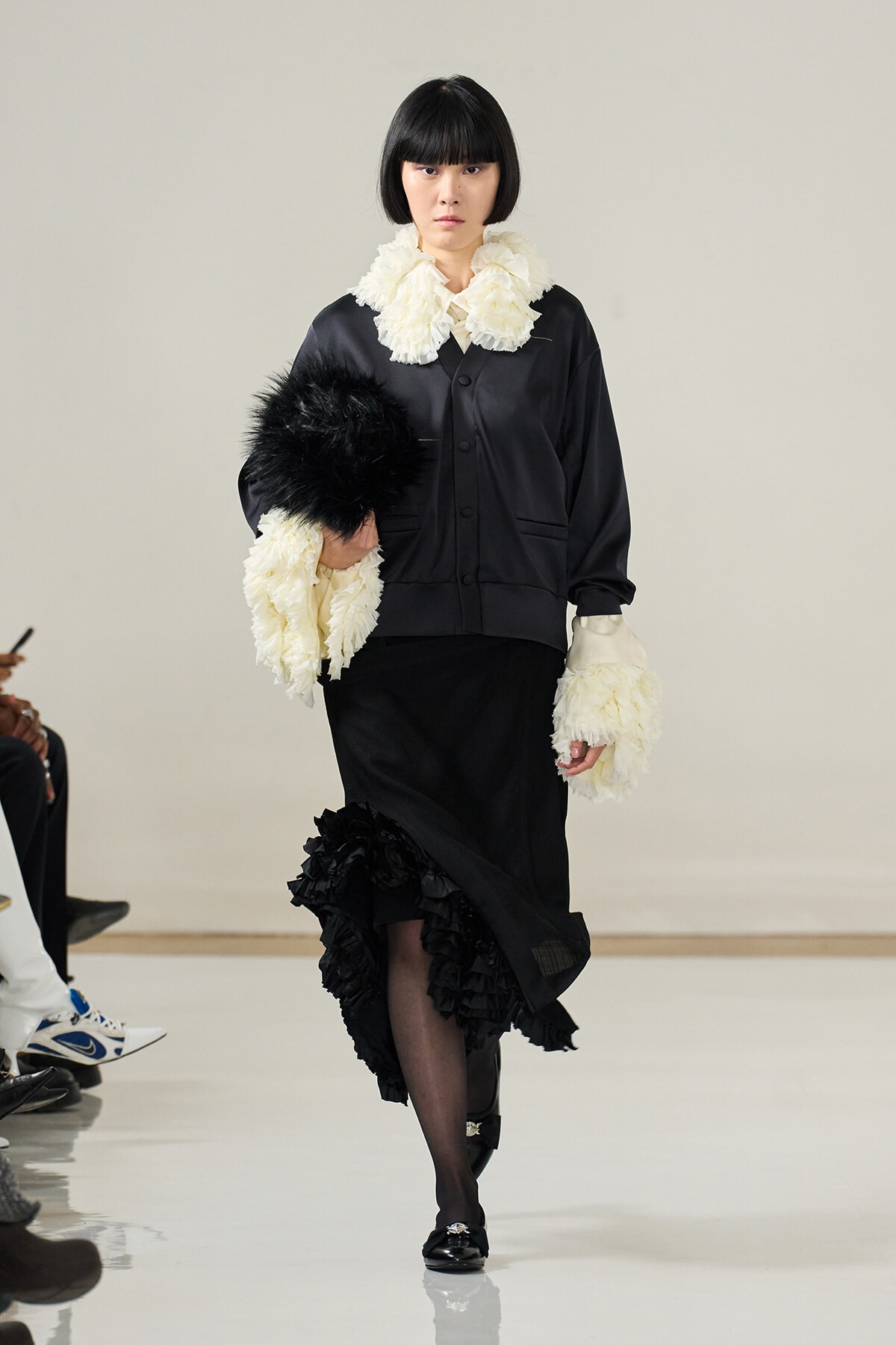 Runway model in a black satin outfit with white ruffled collar and cuffs, carrying a black fur accessory and wearing black heels.