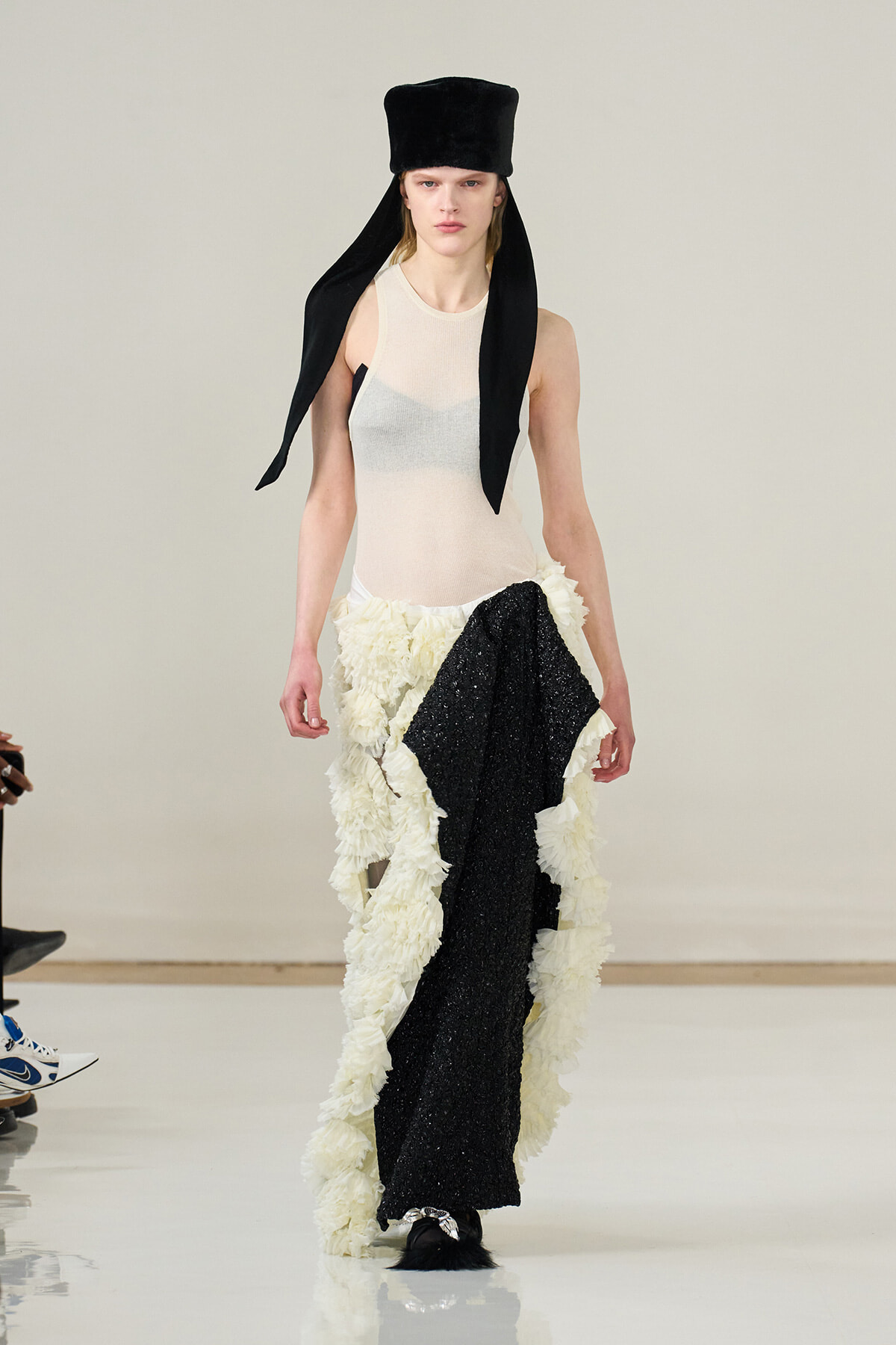 Runway model wearing a tall black hat with long tails, a sheer beige sleeveless top, and a cream ruffled skirt with a black sequined panel.