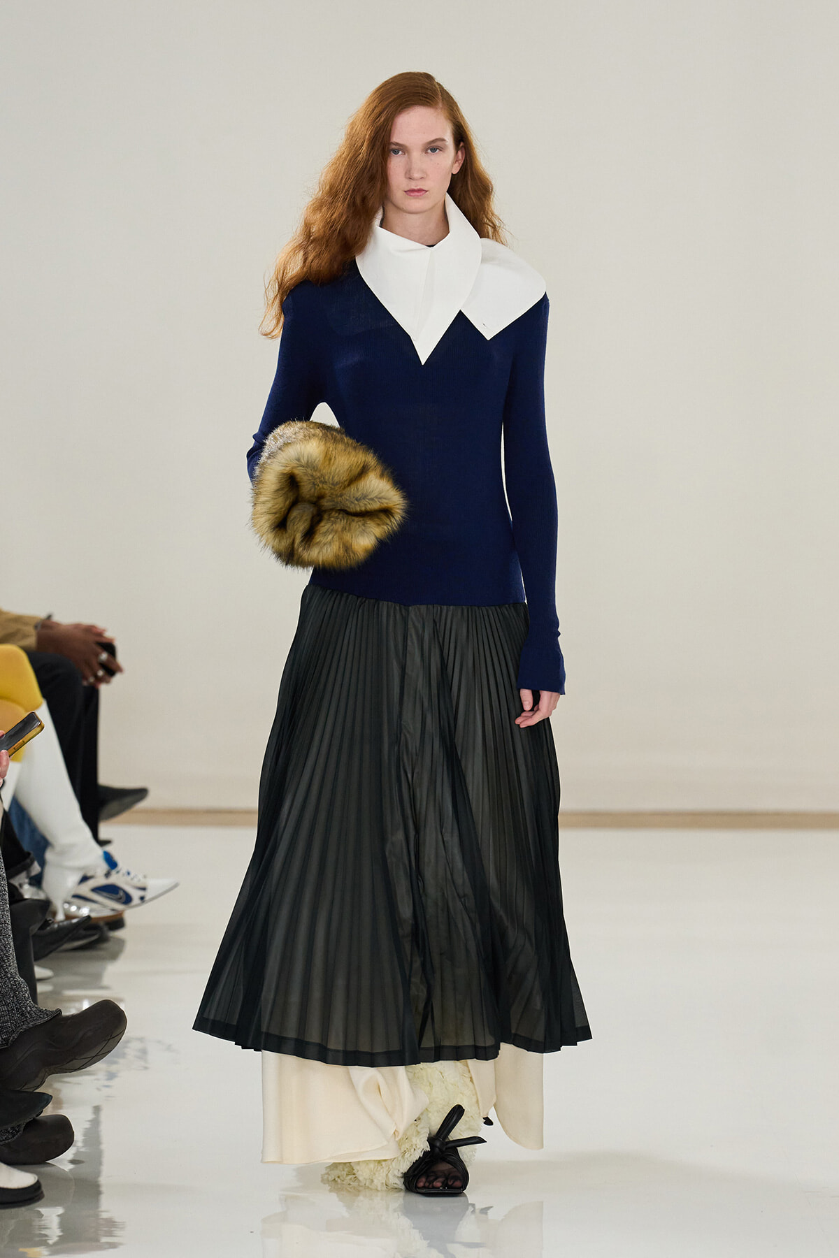 Model walking runway wearing a navy top with a large white collar, paired with a black pleated skirt and a fur clutch.