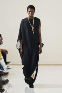 Male model walking a fashion show in a loose black sleeveless cape with floral-lined inner panels and a long pearl necklace.