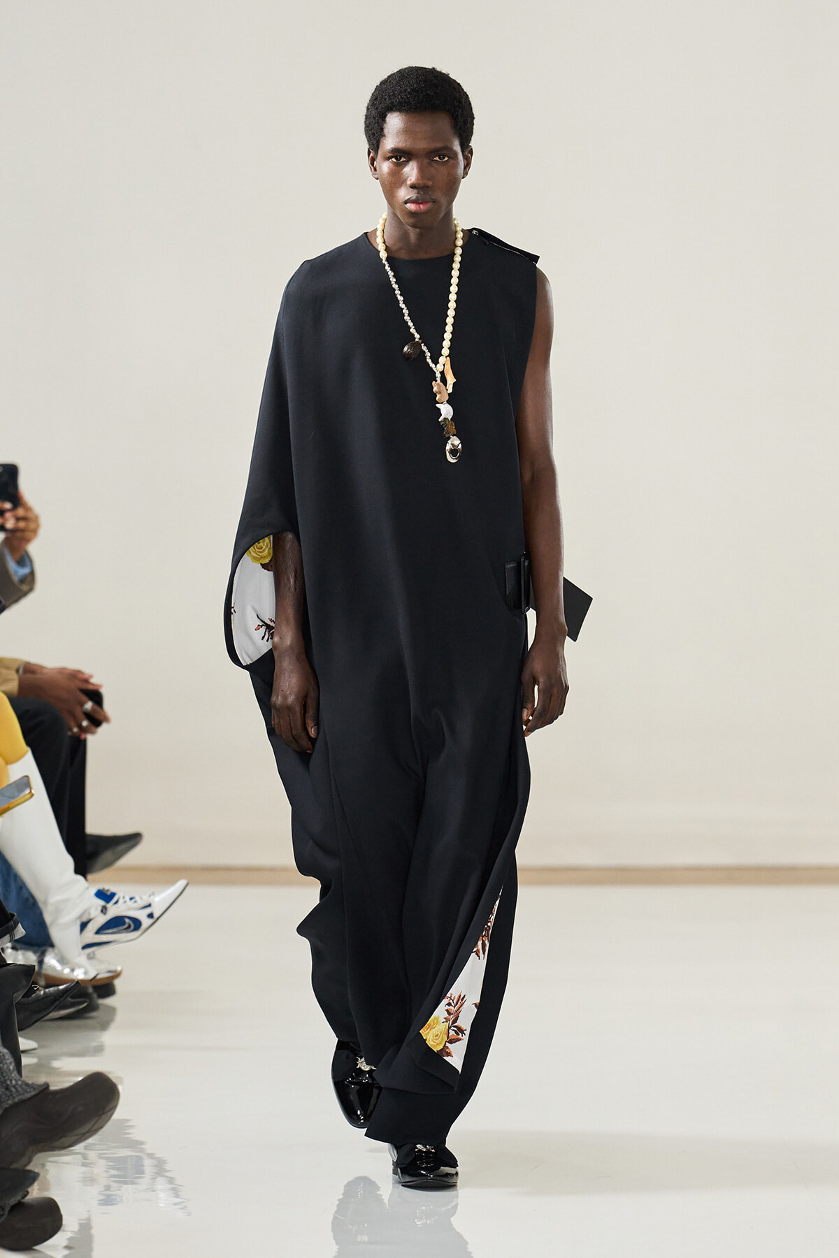 Male model walking a fashion show in a loose black sleeveless cape with floral-lined inner panels and a long pearl necklace.