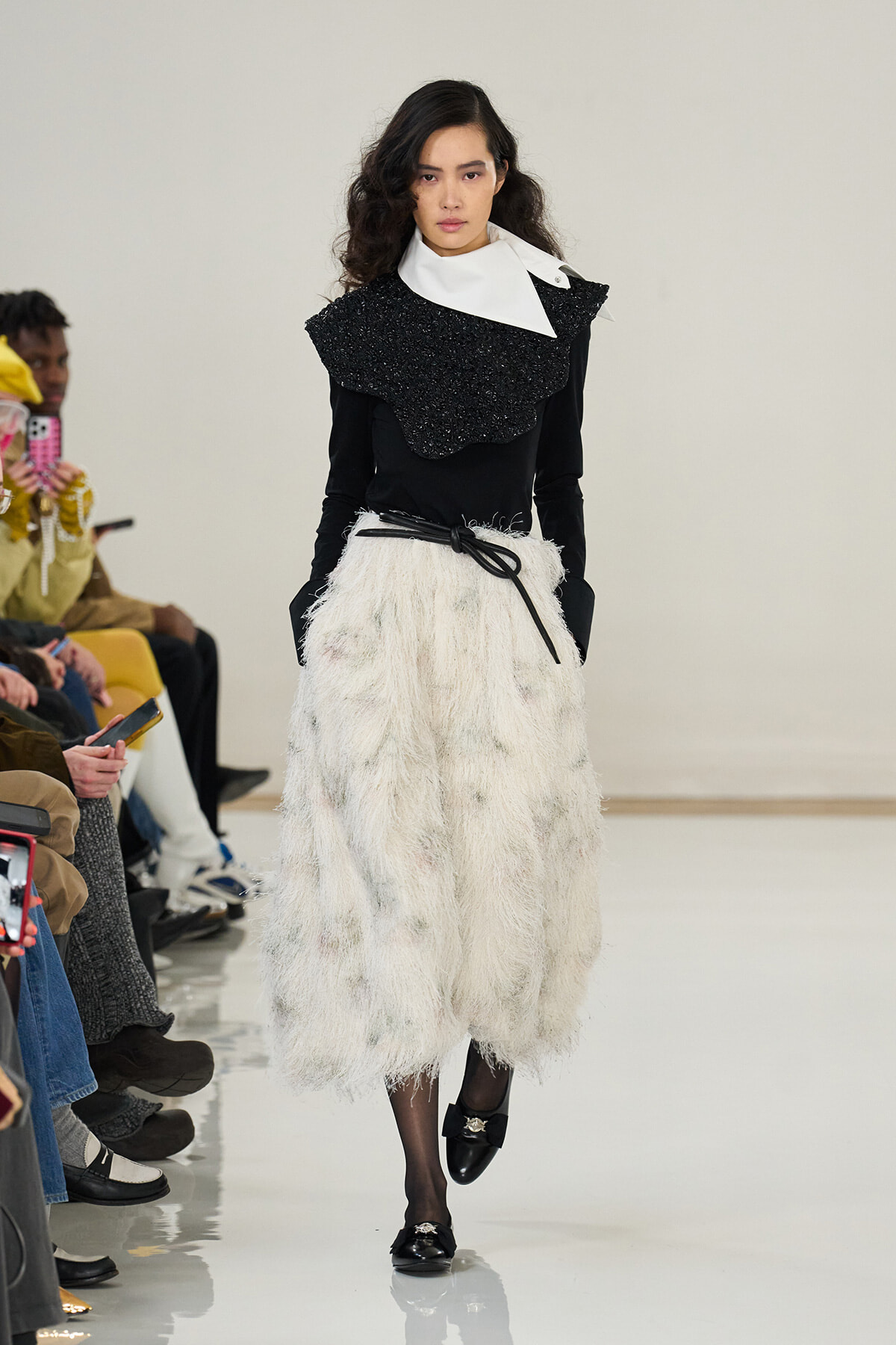 Model walking a runway in a black long-sleeve top with a textured black cape over a white collar, paired with a voluminous white fur-like skirt and a thin black belt.