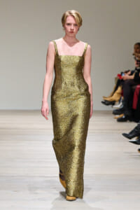 Model walks a gold sequined sleeveless gown down a runway, neutral expression, audience seated at sides.