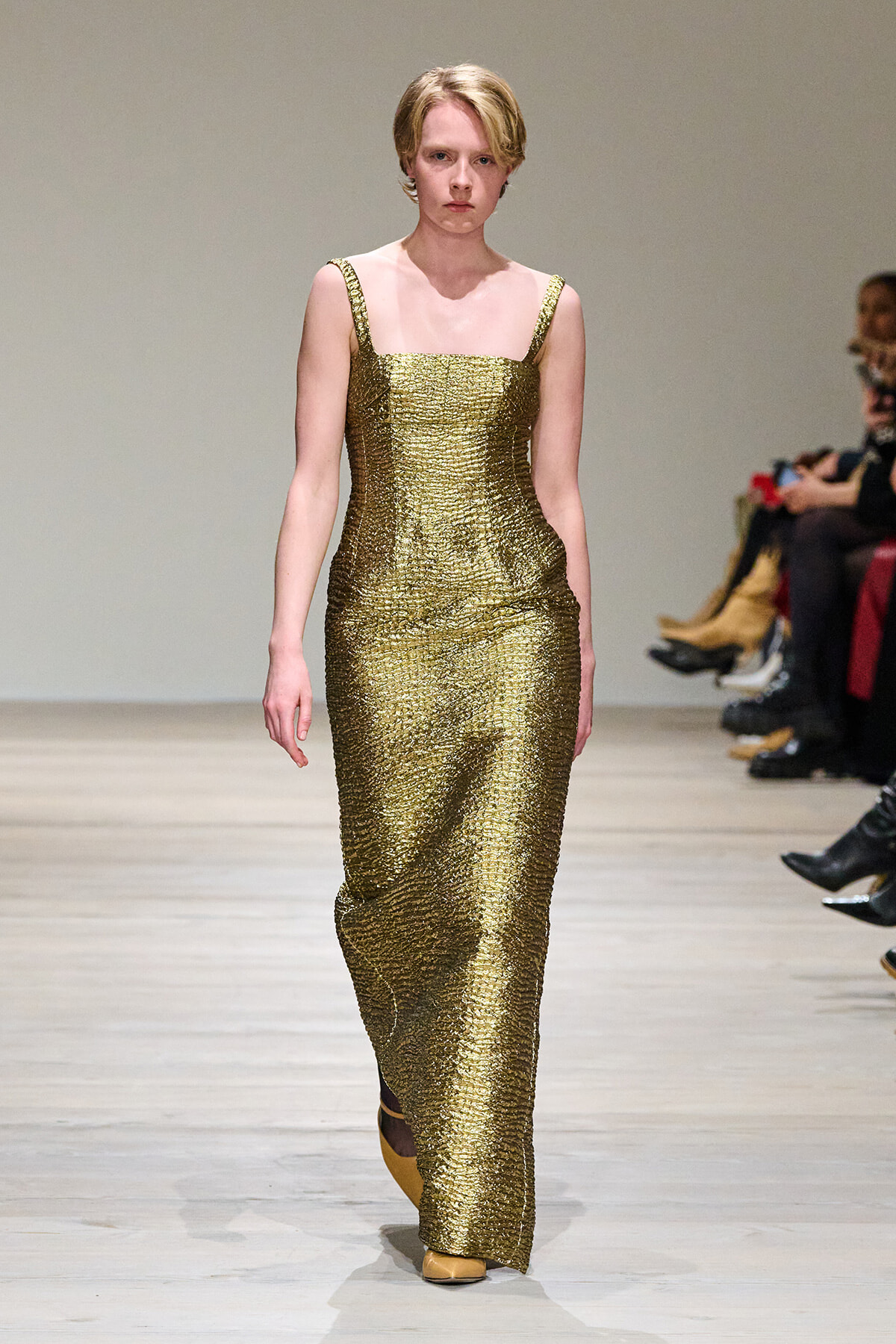Model walks a gold sequined sleeveless gown down a runway, neutral expression, audience seated at sides.