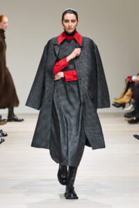 Model walking a fashion runway in a gray check coat with red collar and cuffs, black boots.