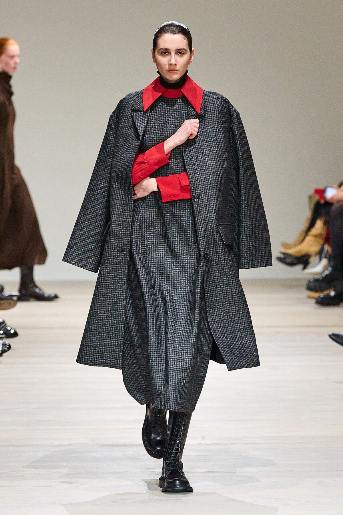 Model walking a fashion runway in a gray check coat with red collar and cuffs, black boots.