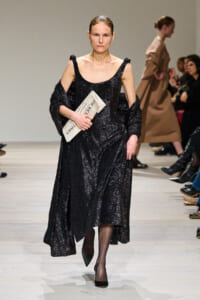 Model walking the runway in a long black sequined dress, holding a folded newspaper under her arm as she stares ahead. The outfit features off-shoulder straps and a dramatic drape.