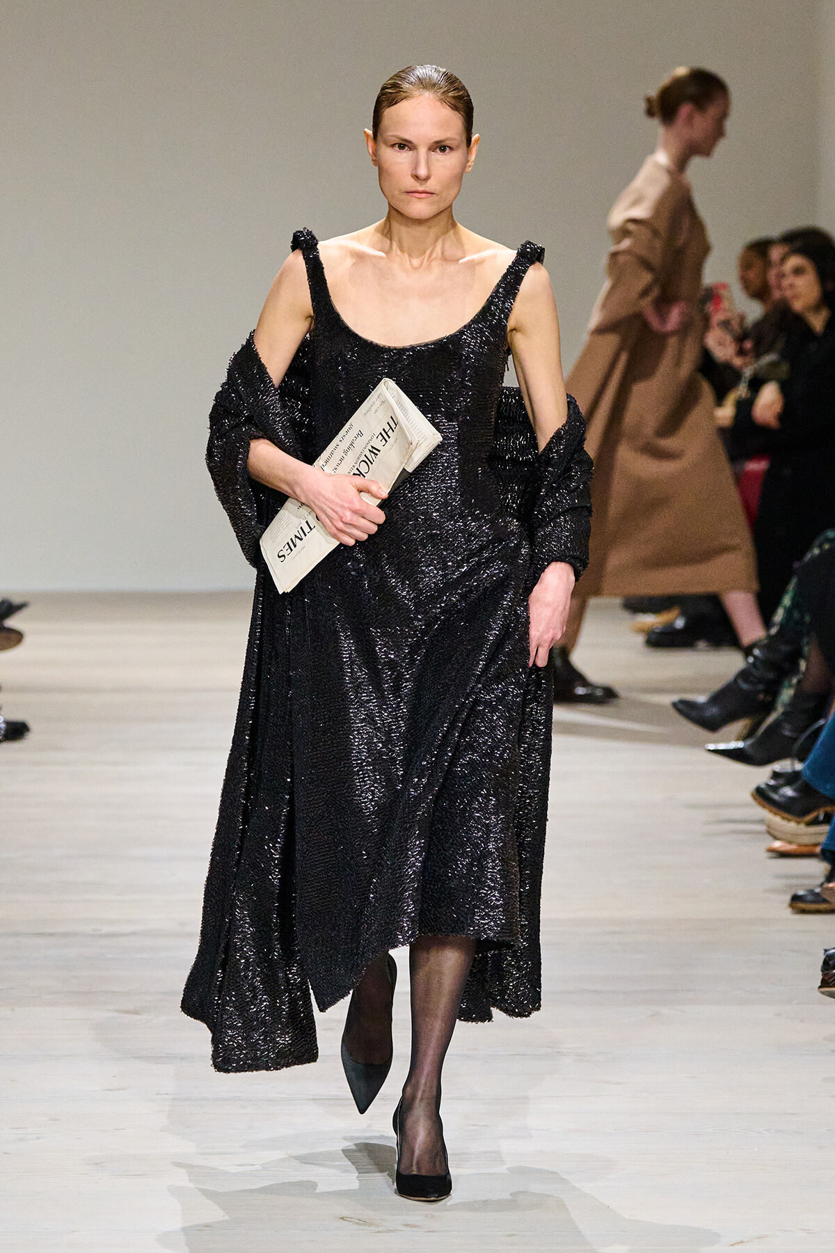 Model walking the runway in a long black sequined dress, holding a folded newspaper under her arm as she stares ahead. The outfit features off-shoulder straps and a dramatic drape.