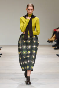 Model walks a runway in a lime knit sweater, black scarf, and a dark green plaid midi skirt with brown lace-up boots.