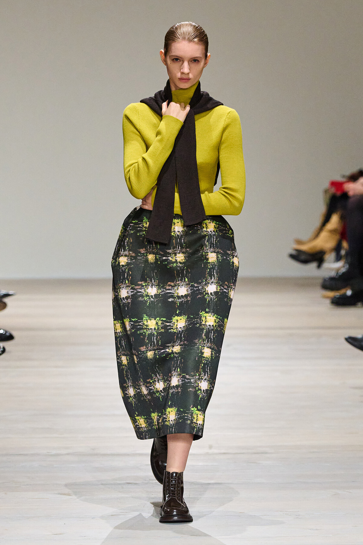 Model walks a runway in a lime knit sweater, black scarf, and a dark green plaid midi skirt with brown lace-up boots.