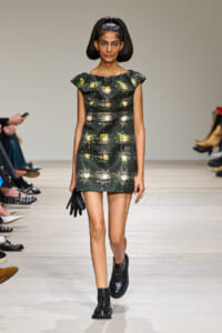 Model on a runway wearing a dark green abstract print mini dress with cap sleeves, black ankle boots, and a black glove clutch in her left hand