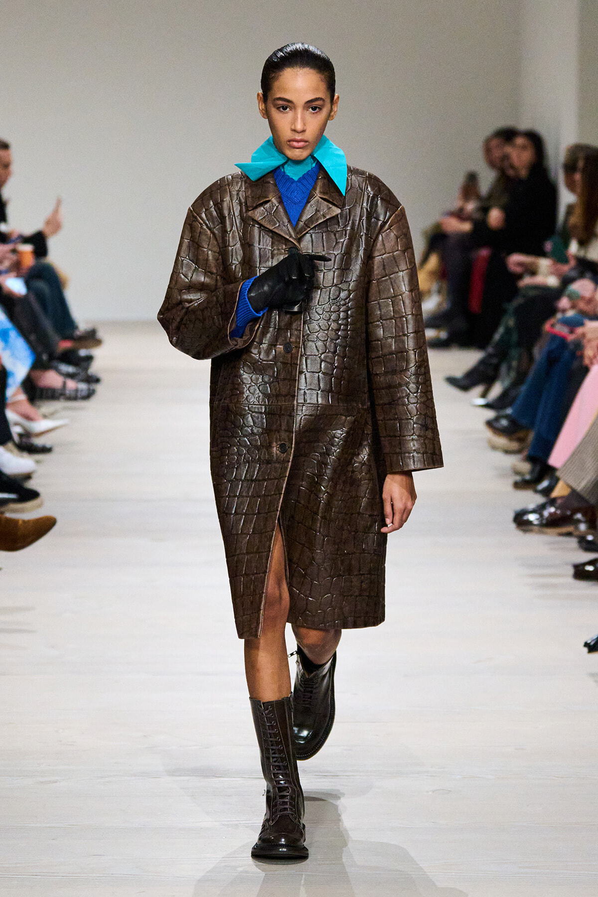 Model walks the runway in a brown crocodile-embossed coat over a blue sweater and teal scarf, with black gloves and tall lace-up boots.