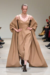 Model walking the runway in a tan, structured dress with a matching voluminous cape draped over shoulders.