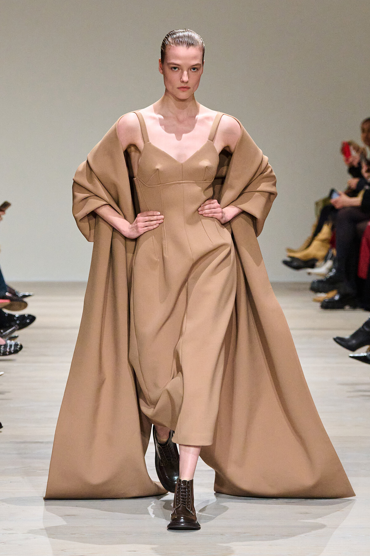 Model walking the runway in a tan, structured dress with a matching voluminous cape draped over shoulders.