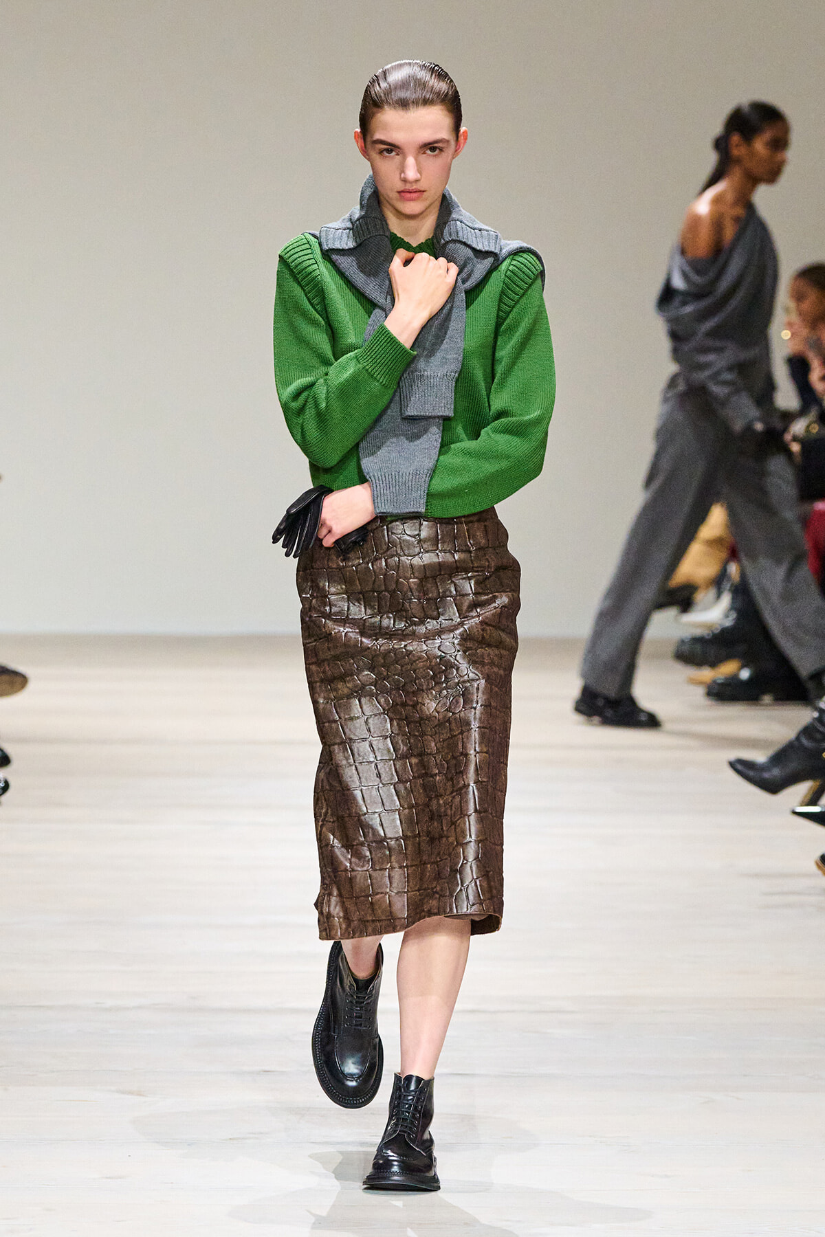 Model walking the runway in a green knit sweater, grey scarf, and a brown crocodile-embossed midi skirt with black boots.