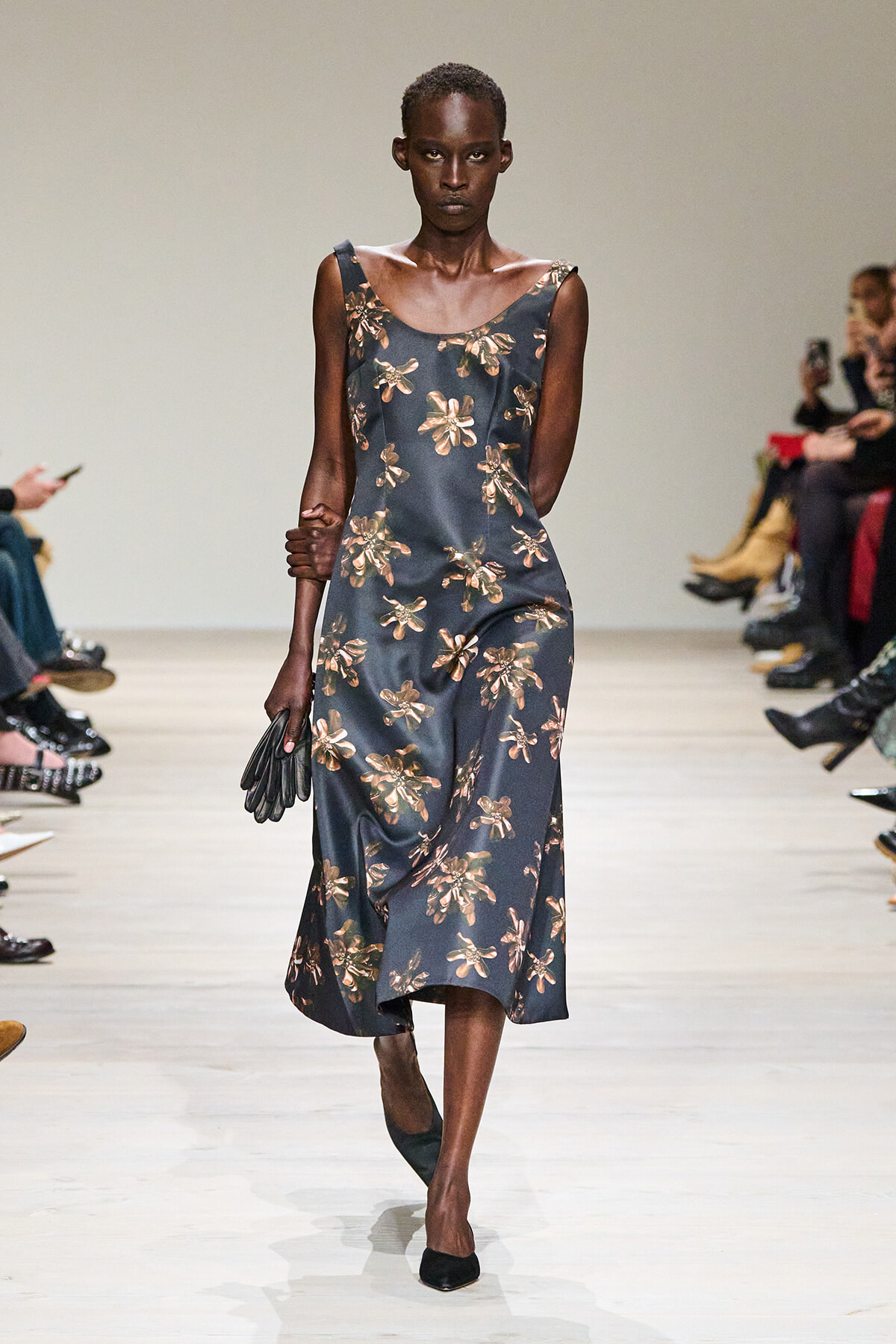 Model walks the runway in a dark blue floral midi dress, holding black gloves, with a neutral backstage audience in the background.