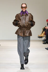 Model on a runway wearing a brown crocodile-textured jacket with a purple collar, gray wide-leg pants, black gloves, and sunglasses, facing forward