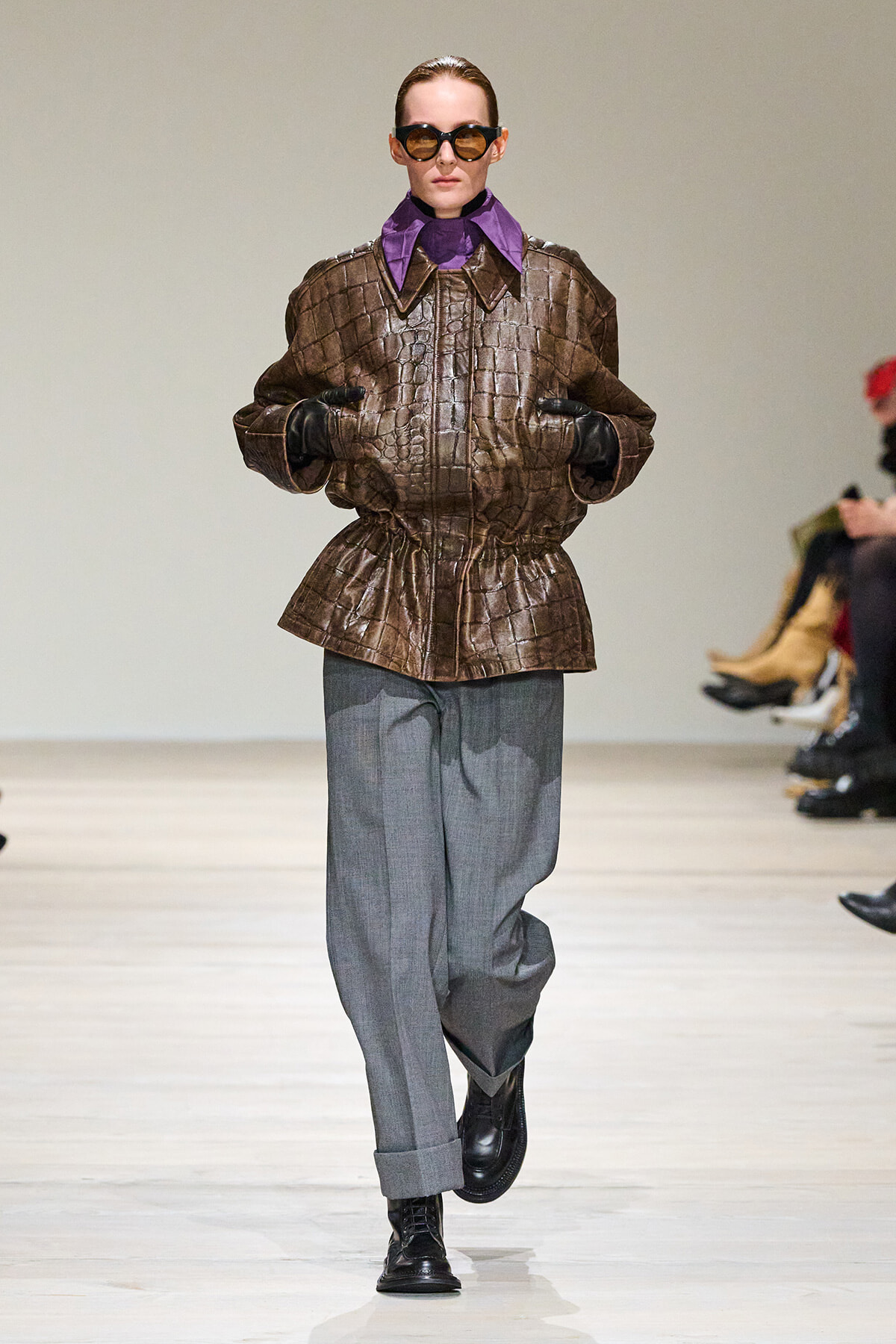 Model on a runway wearing a brown crocodile-textured jacket with a purple collar, gray wide-leg pants, black gloves, and sunglasses, facing forward