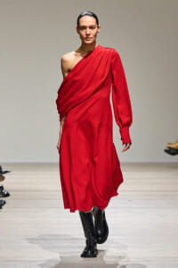 Model wearing a red one-shoulder draped dress and black knee-high boots walking a runway.
