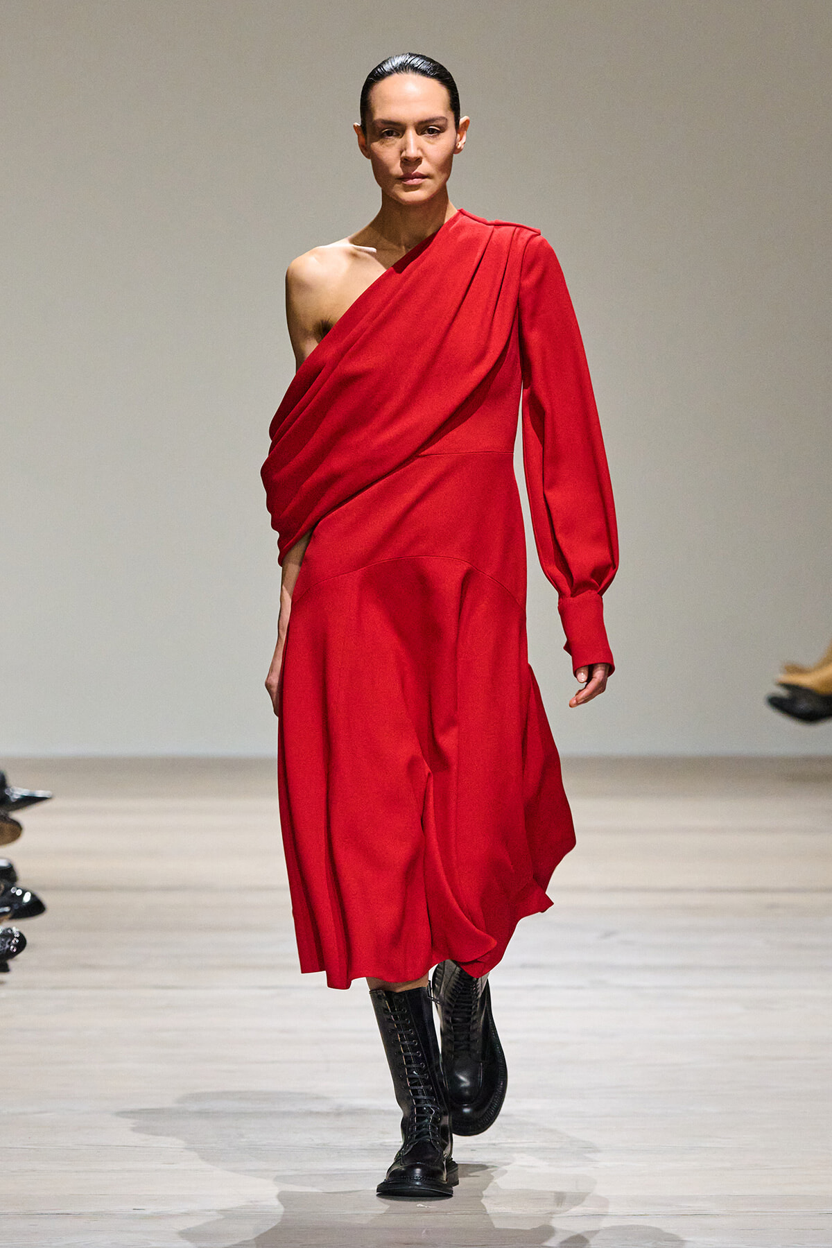 Model wearing a red one-shoulder draped dress and black knee-high boots walking a runway.