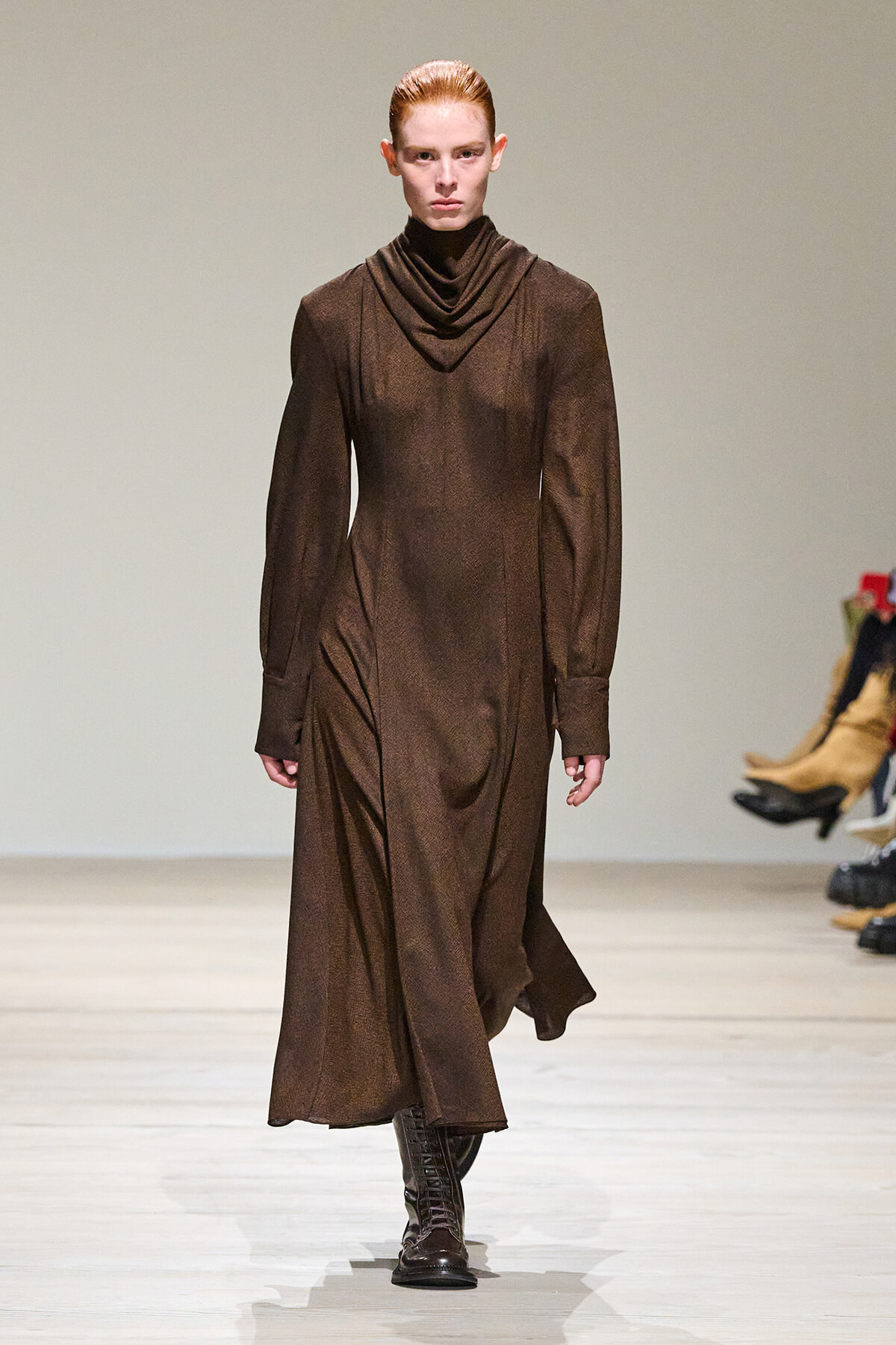 Model walking runway in a long brown, draped-neck dress with full sleeves and lace-up boots.
