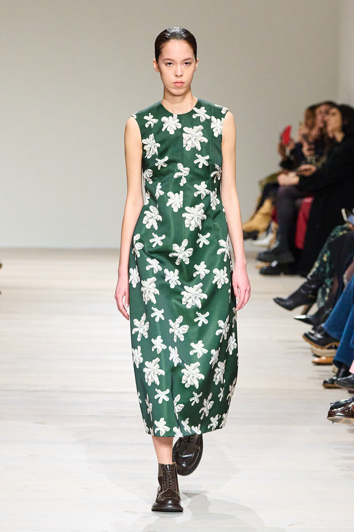 Model walking runway in a sleeveless dark green midi dress with white floral print and dark ankle boots.